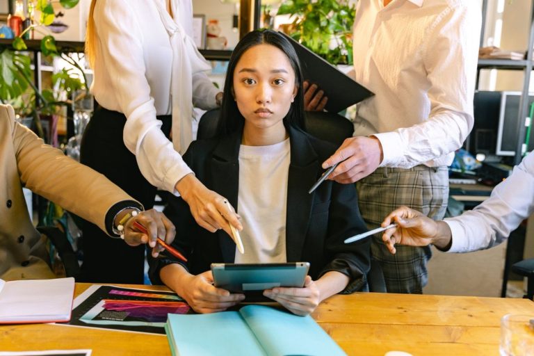 Une manager assise devant une tablette est entourée de plusieurs collaborateurs qui pointent leurs stylos vers elle, illustrant la pression managériale et le manque de légitimité dans la prise de décision.